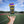 A translucent gray shaker bottle with a purple and yellow lid is floating above an open hand on an outdoor sports field under a blue sky. The bottle has 'DNA SPORTS' printed in yellow on the front.